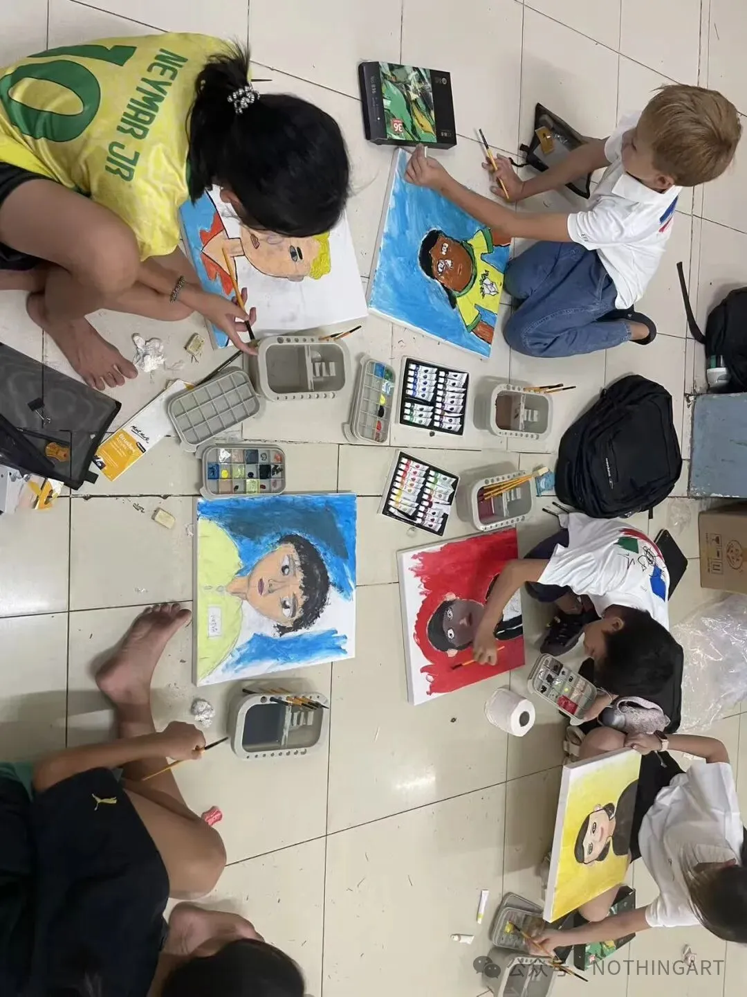 An overhead view of participants painting colourful portraits, with canvases, brushes and paint sets arranged on the floor.